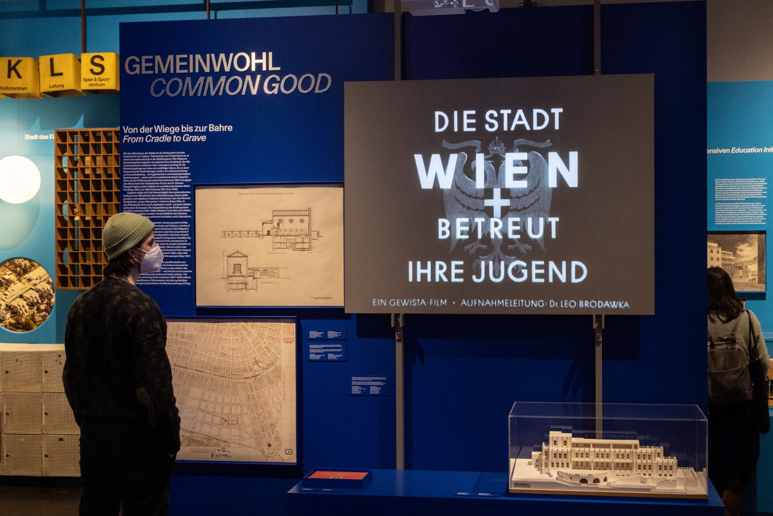 blaue Ausstellungswand mit Screen "Die Stadt Wien betreut ihre Jugend" davor steht ein Mann mit Haube