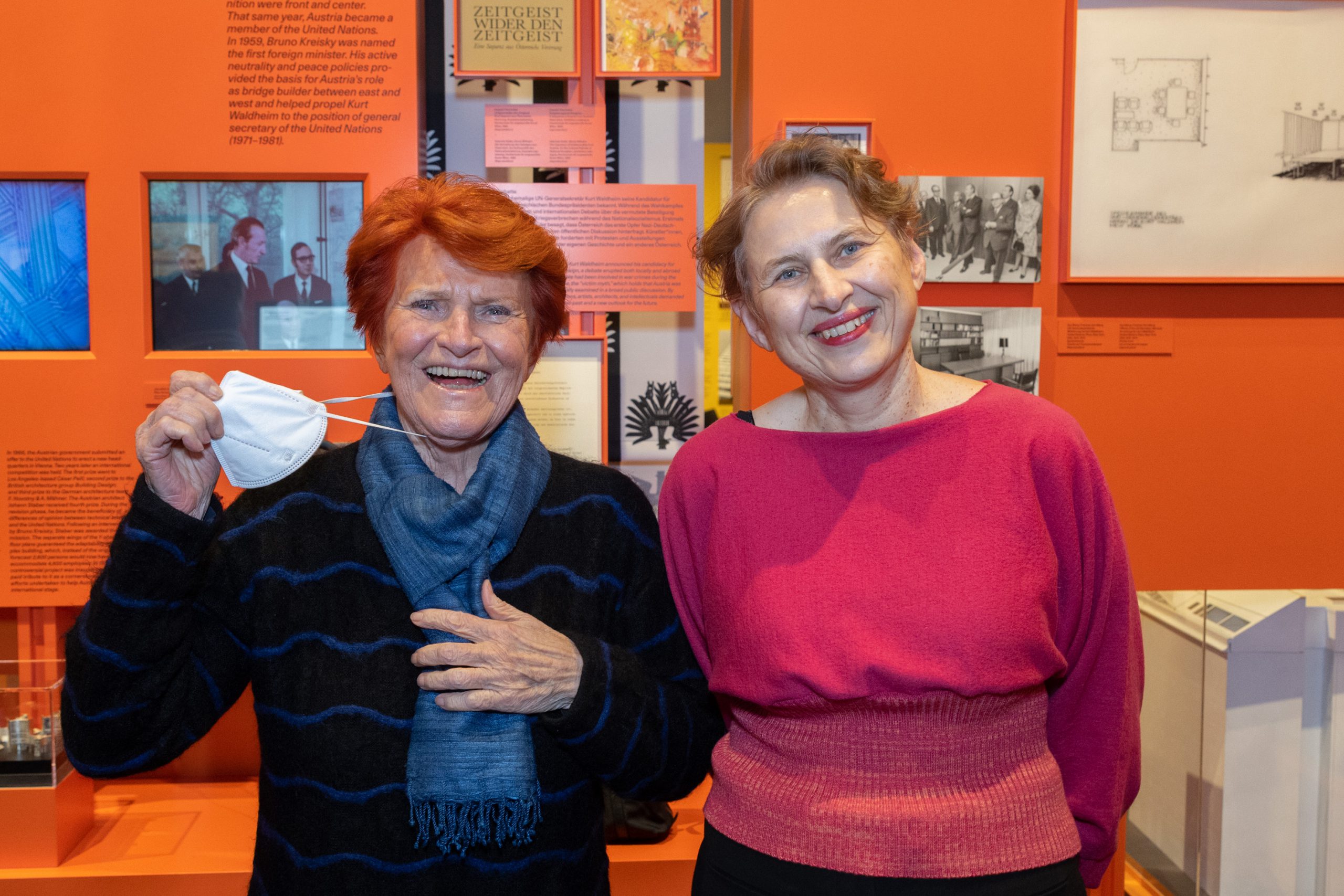 2 lachende Frauen, eine davon mit einer Maske, die andere mit pinken Pulli vor oranger Ausstellungswand