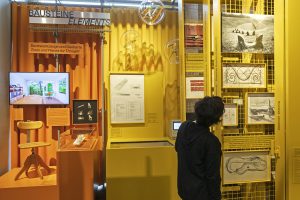 Person schwarz gekleidet vor gelb-oranger Ausstellungswand