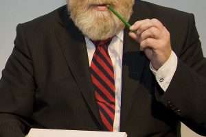 A man with a striped tie, holding a pad and a green pencil in his mouth