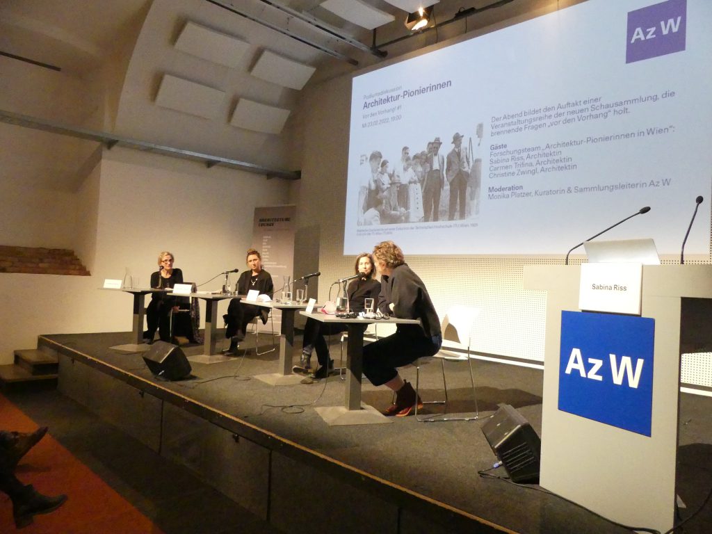 4 darkly dressed women sitting on a podium with microphones