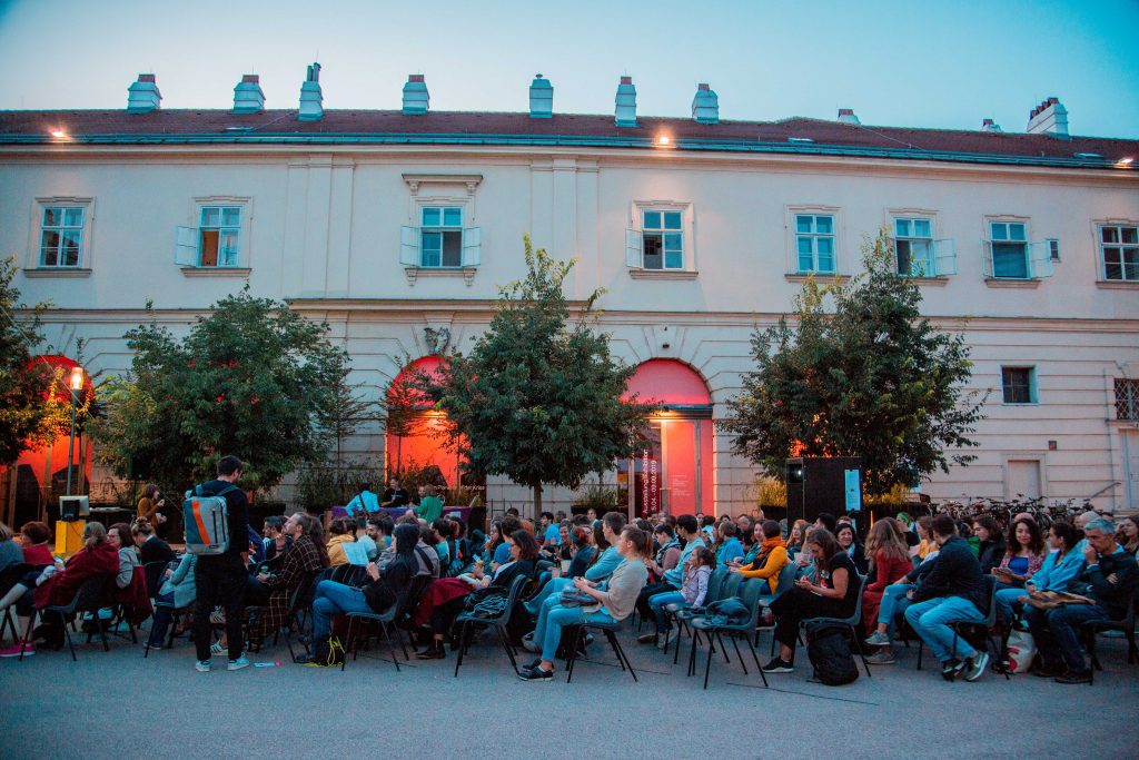 many people sitting on armchairs in a courtyard, behind them 3 trees and an old building