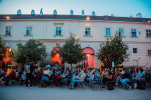 many people sitting on armchairs in a courtyard, behind them 3 trees and an old building