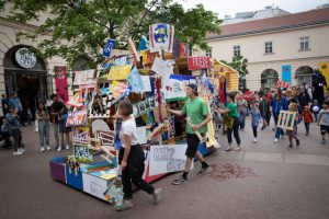 Viele Menschen in einem Innenhof, in der Mitte steht ein großes buntes Objekt aus Tafeln, Schachteln