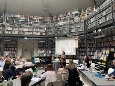 Several people in a room with many bookshelves sitting at tables, one person standing in front