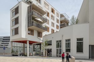 3-4 geschossiges Wohnhaus mit Balkonen und davor Platz aus verschiedenen Steinen