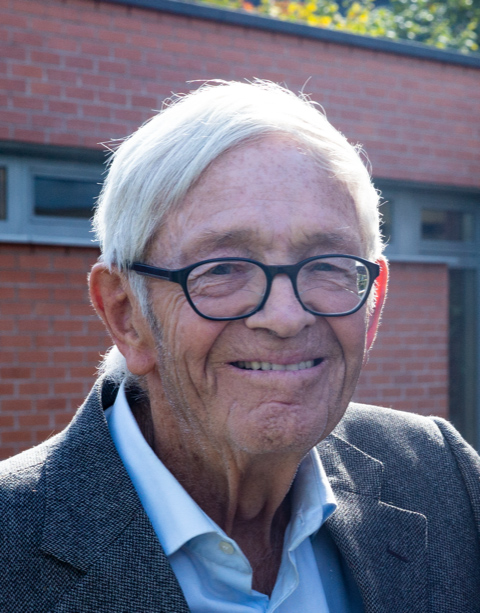 alter weißhaariger Mann mit schwarzer Brille vor einer Ziegelwand