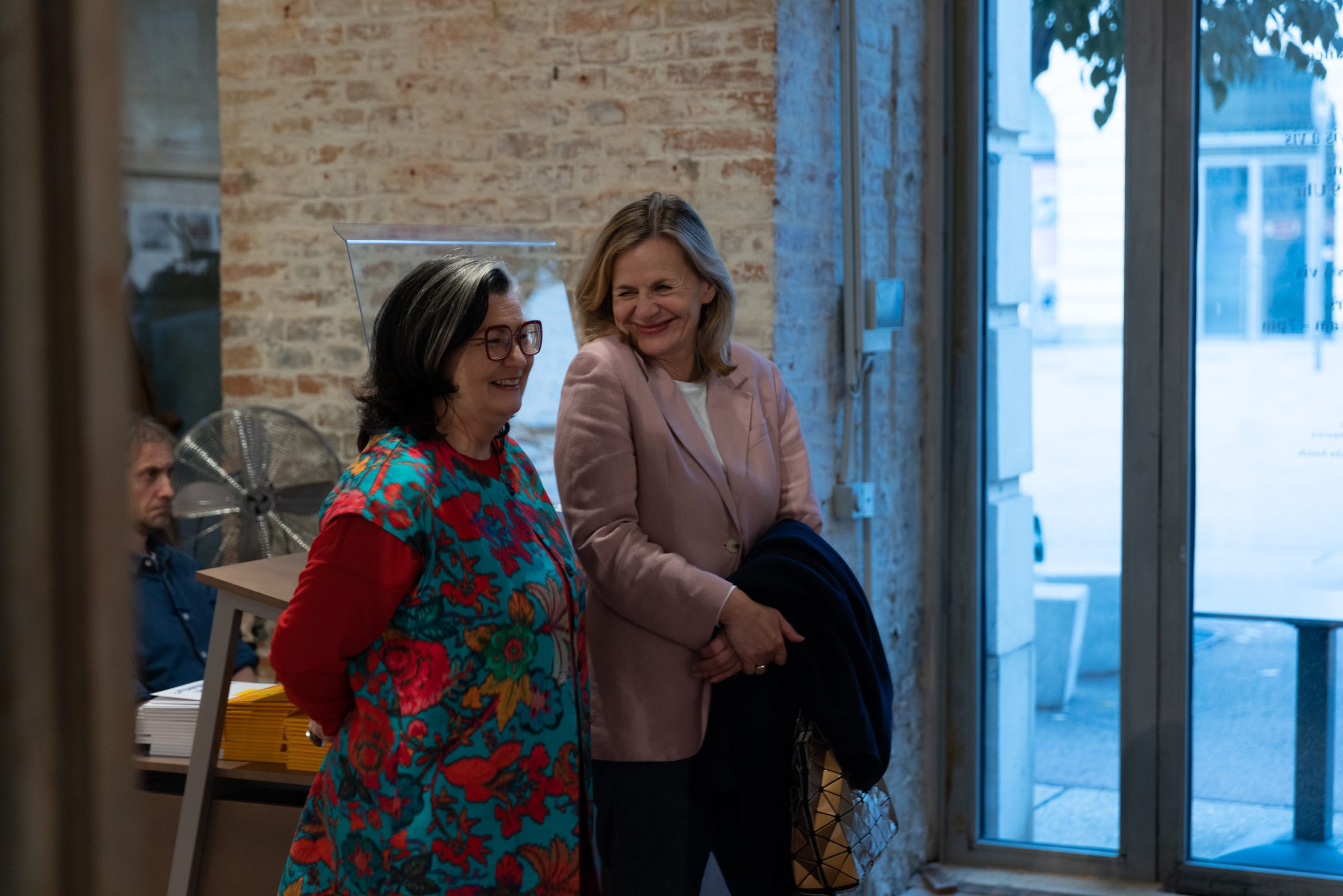 2 women laughing, one of them wears glasses, behind them is a fan