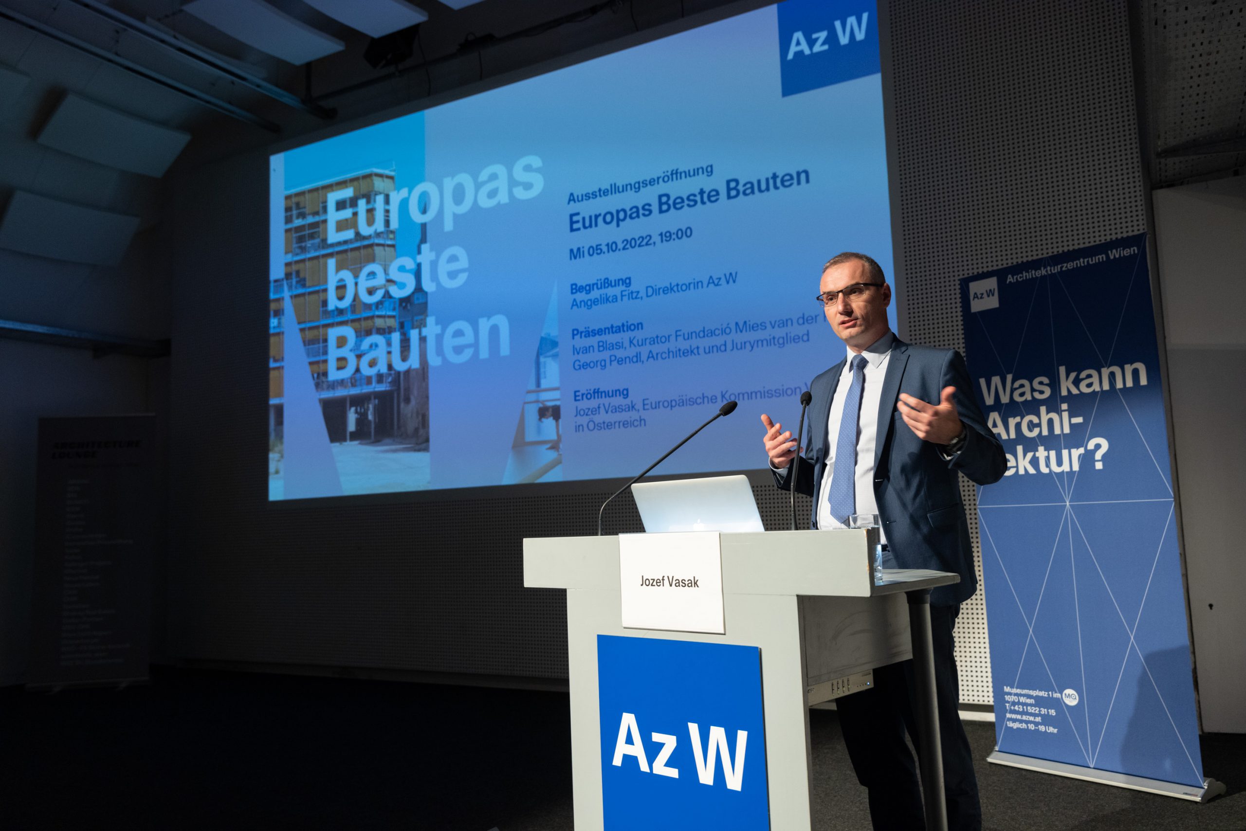 Man with glasses stands behind a desk, behind him a screen with the words "Europe's best buildings"