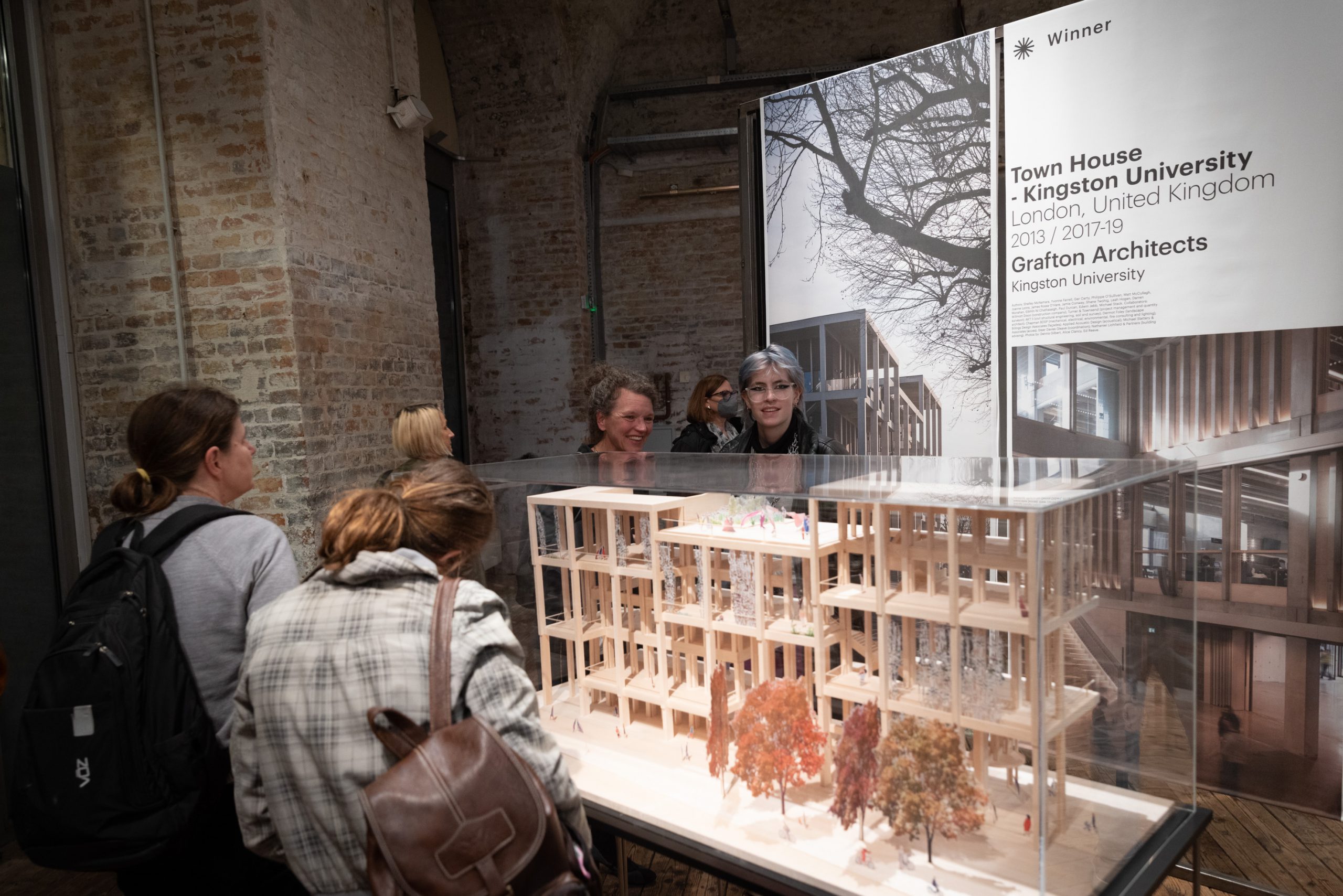 Large wooden architectural model under plexiglass cube, in front of and behind it some people