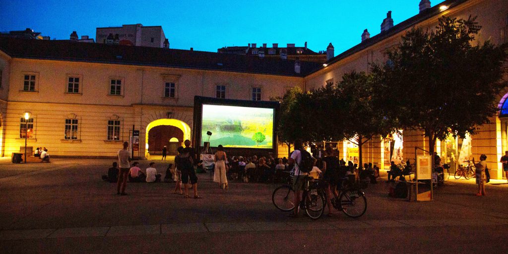 Canvas in a courtyard at dusk with people standing and sitting in front of it