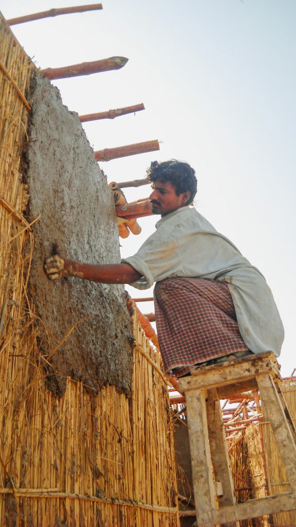 Mann hockt auf einem hohen Schemel und verputzt eine Bambuswand