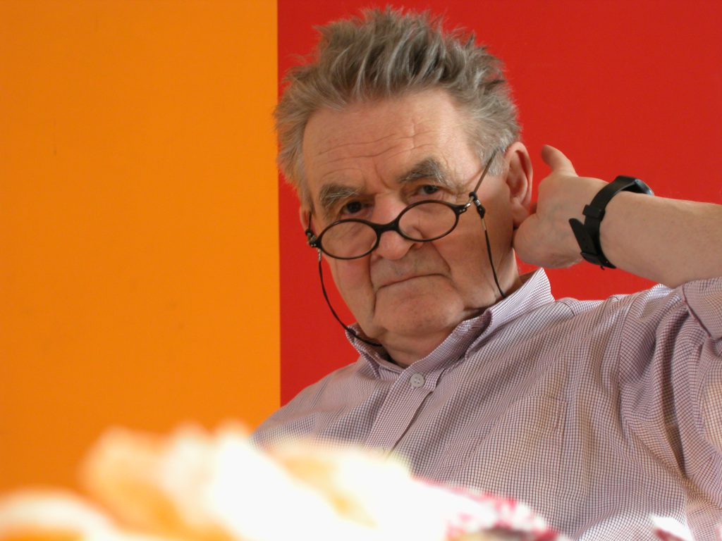älterer Mann vor einer rot-orangen Wand mit runder Brille