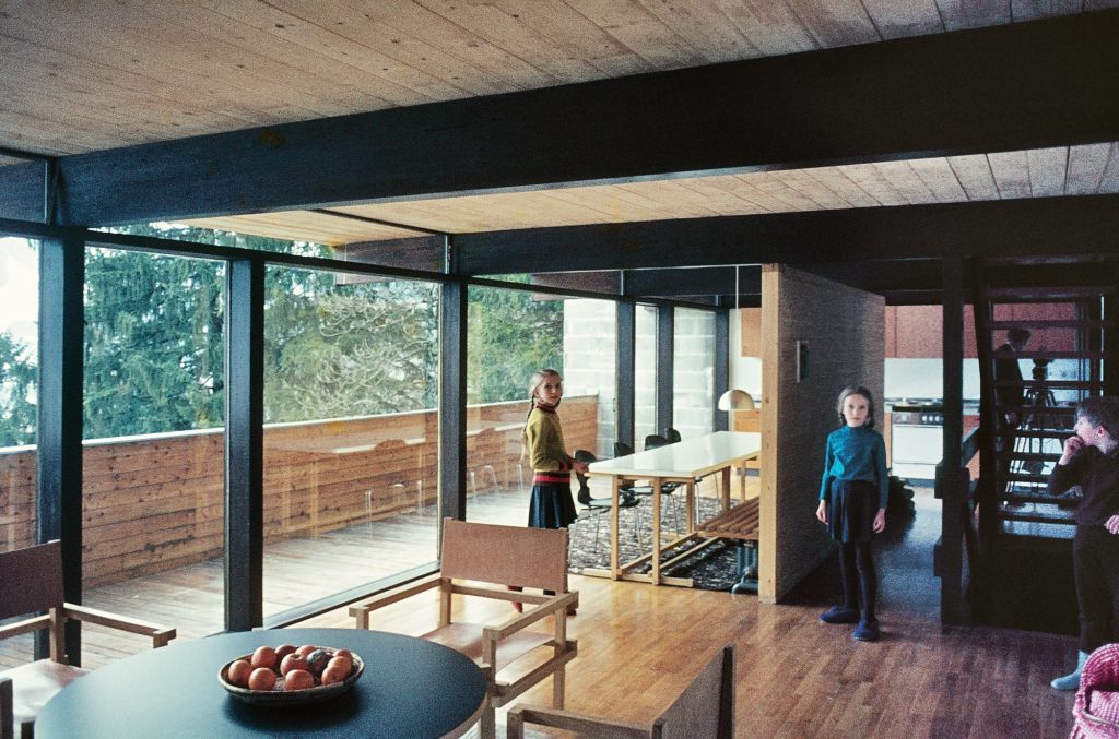 Interior with long glass façade and round table with fruit on it and 3 children in the background