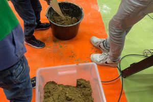 Red-green ground, on it a few people stirring mud in a bucket