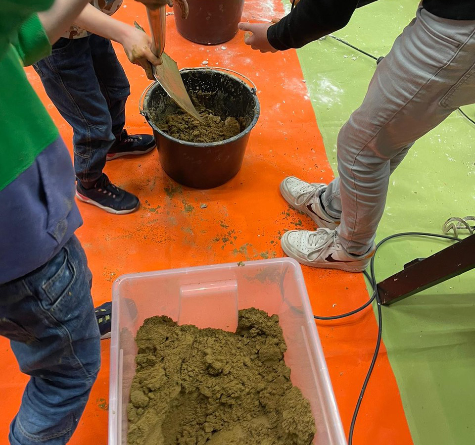 rot-grüner Boden, darauf ein paar Menschen, die in einem Kübel Matsch verrühren