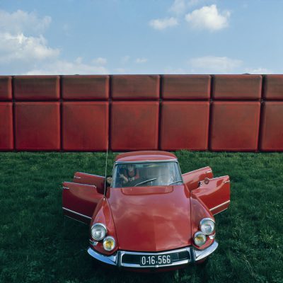 Red car with open doors stands on a meadow with a red-brown wall behind it