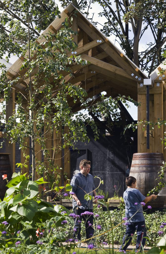 2 Menschen in einem Garten mit Hütte im Hintergrund, Bub hat Gartenschlauch in der Hand