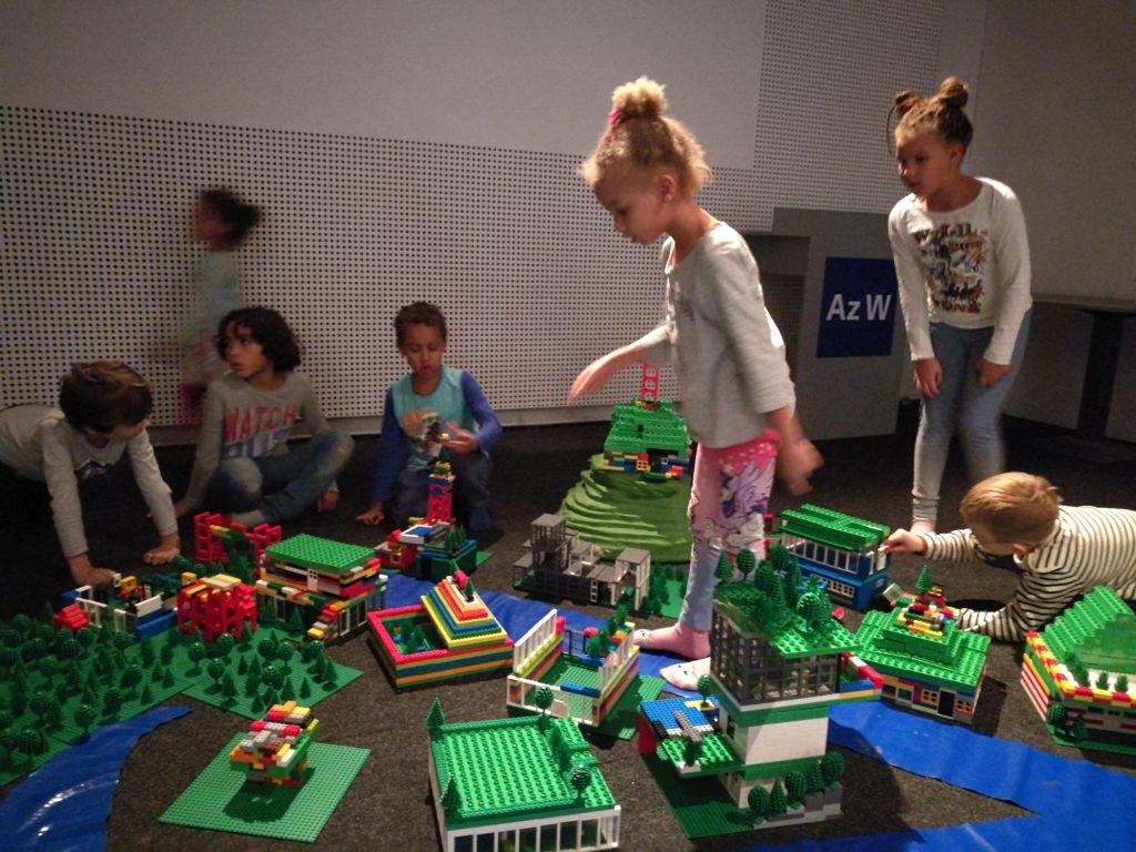 Some children playing with lots of Lego, behind them a wall with many small holes.