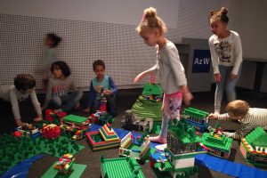 Some children playing with lots of Lego, behind them a wall with many small holes.