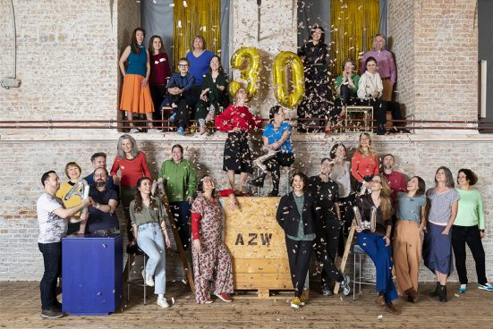 Many colourfully dressed people in front of a brick wall, confetti flying through the air and the number 30 stuck to the wall