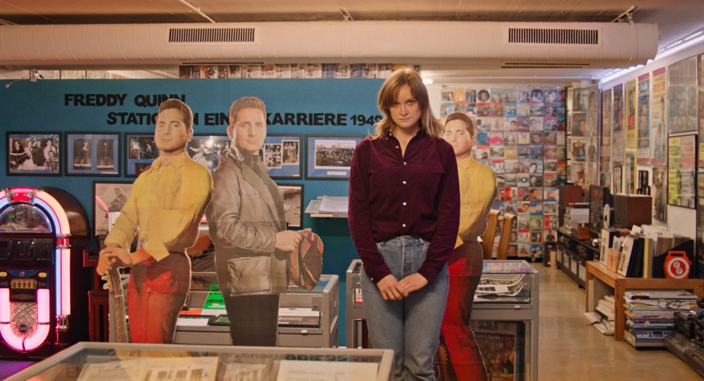 A woman in jeans and a dark red blouse stands in a shop, behind her are cardboard figures