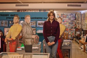 A woman in jeans and a dark red blouse stands in a shop, behind her are cardboard figures