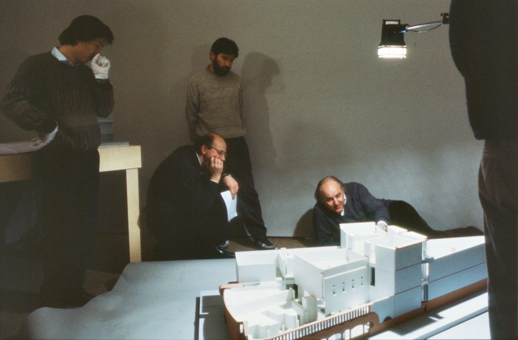 4 men standing and lying in front of a large architectural model