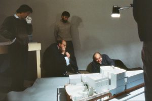 4 men standing and lying in front of a large architectural model