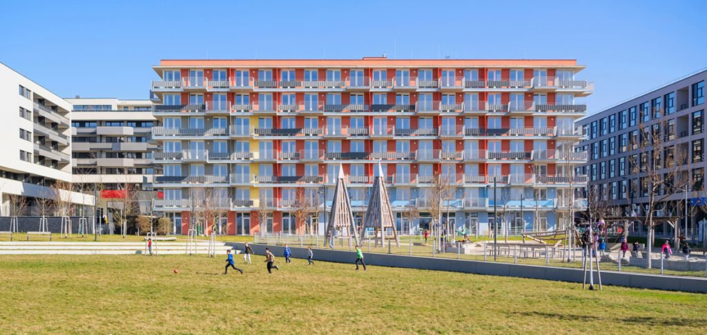 rot farbenes Gebäude mit vielen Balkonen, davor großer Spielplatz mit Wiese