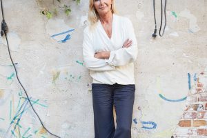 a woman with blond hair stands with folded arms in front of a wall