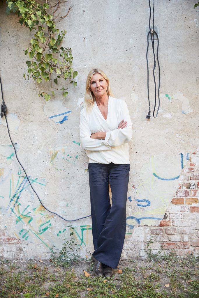 a woman with blond hair stands with folded arms in front of a wall