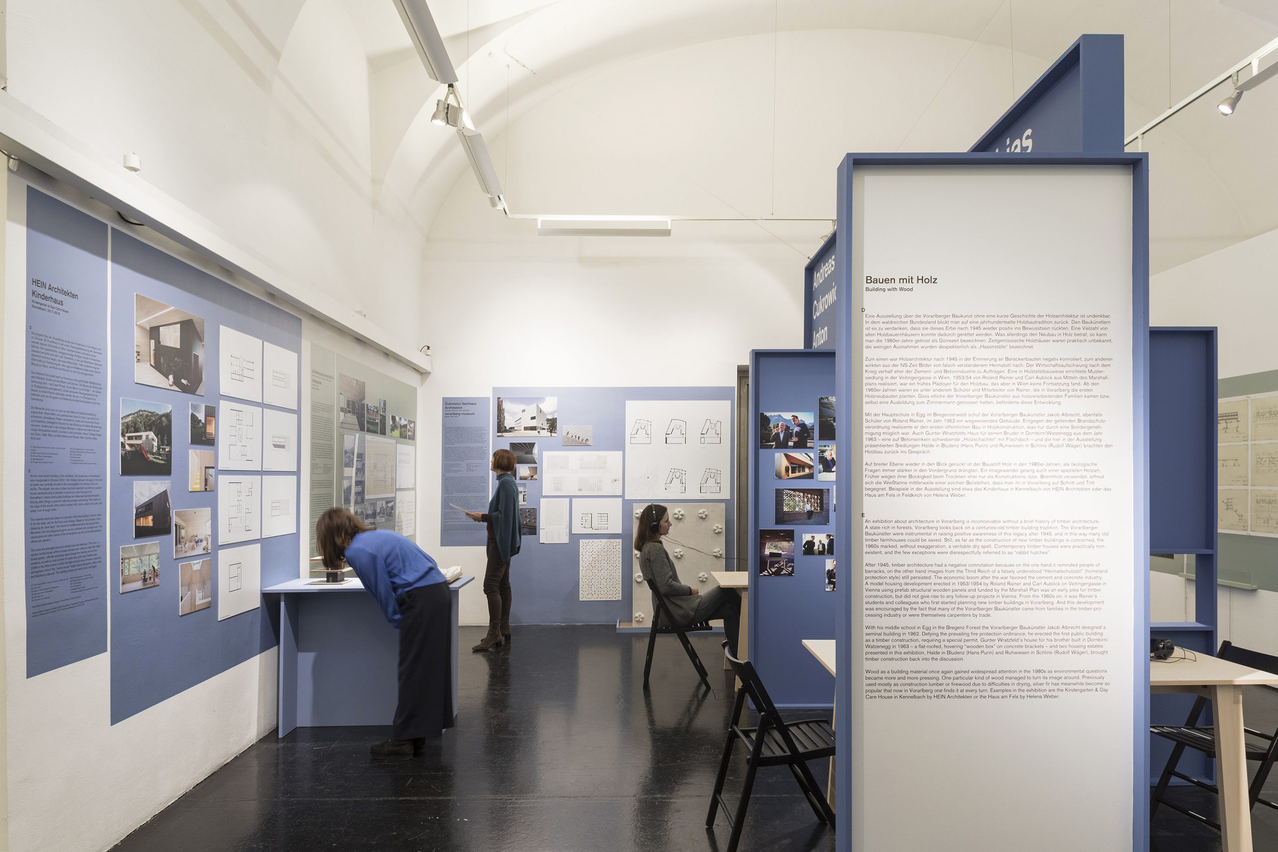 Ausstellungsraum mit blauen Tafeln und Wänden, 2 Personen stehen, 1 Person sitzt mit Kopfhörern