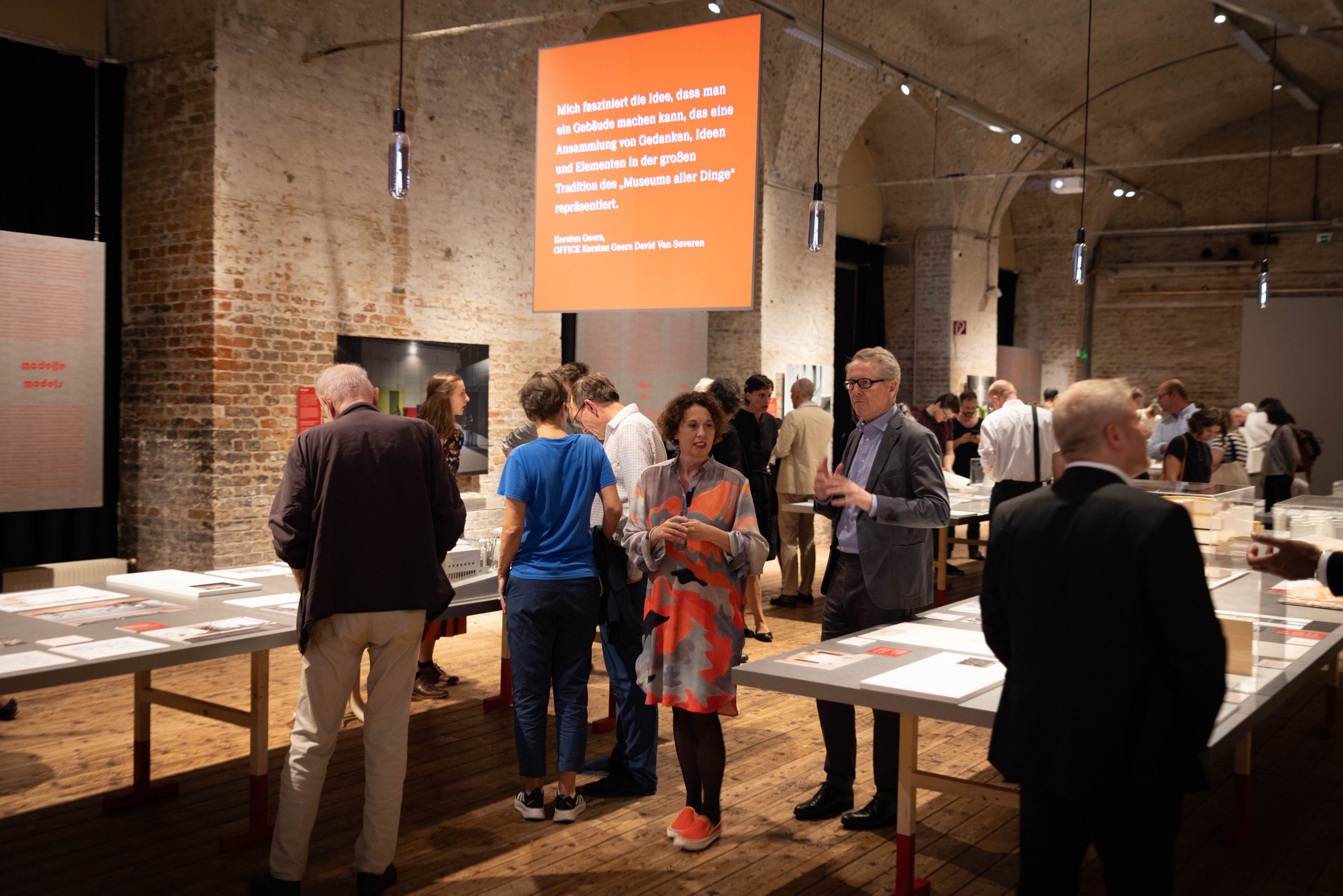 People in an exhibition, from the ceiling hangs a screen in orange colour with white writing