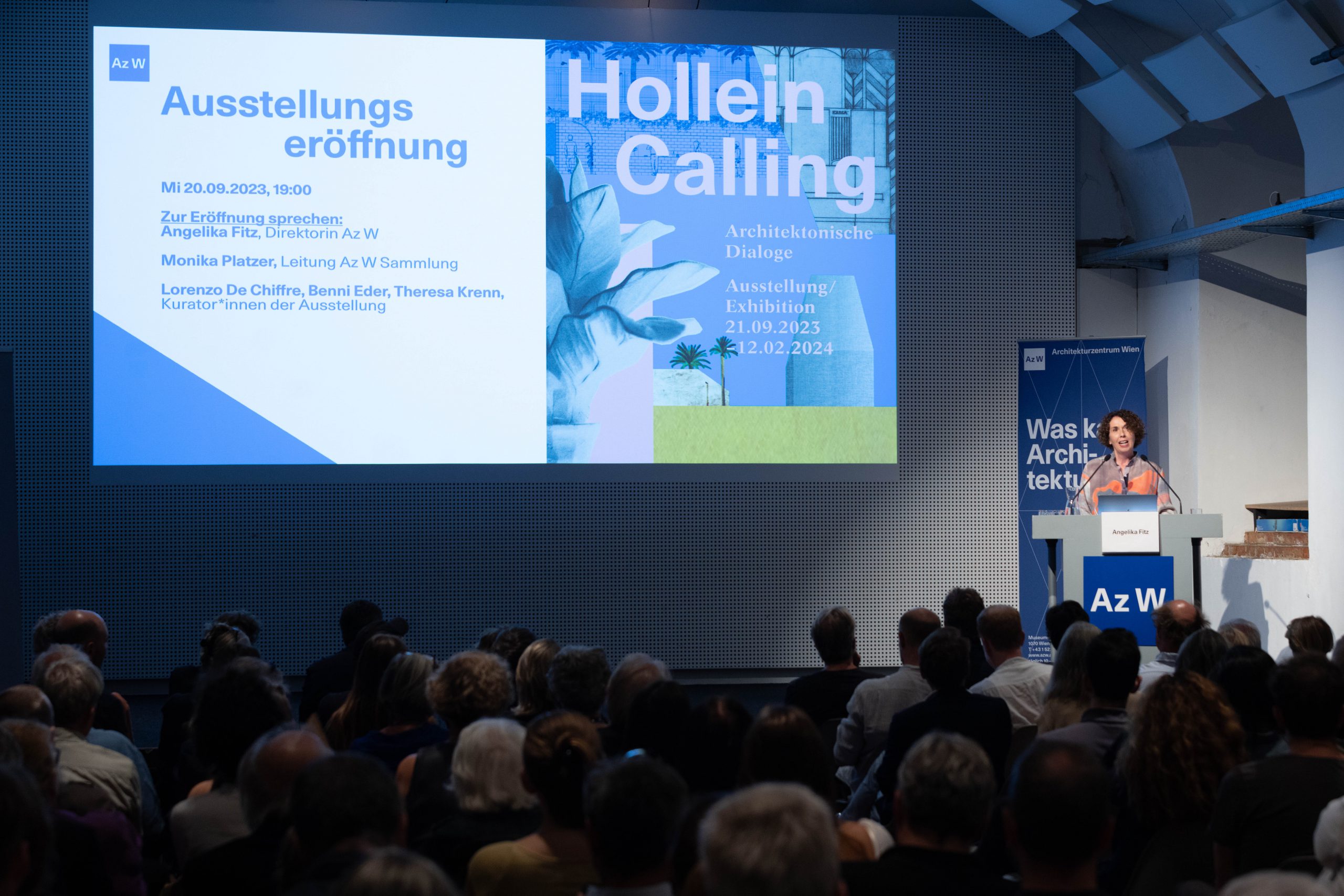 Filled lecture hall with large screen and woman standing at the lectern