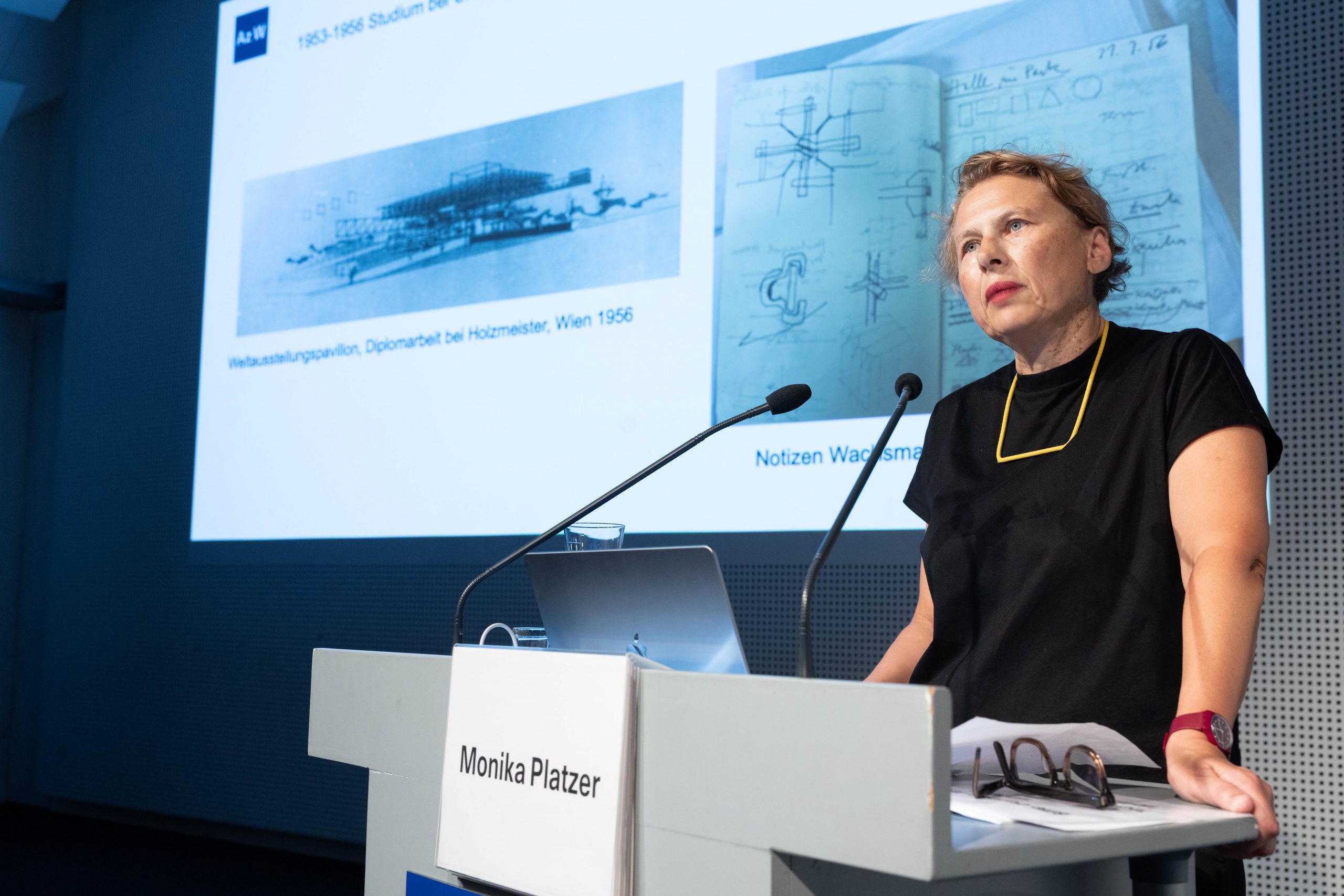 Woman with black shirt and square yellow chain stands at a lectern