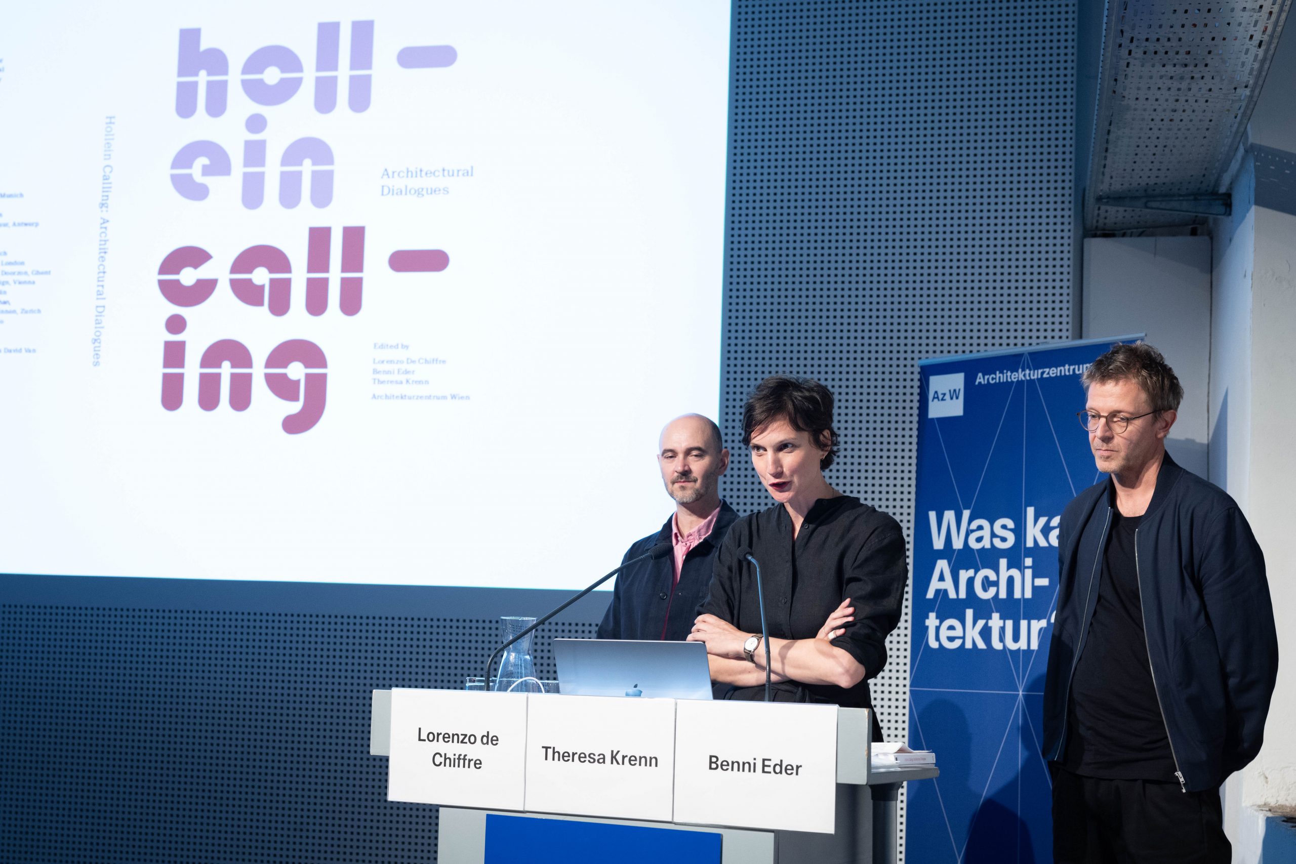 3 people dressed in black - 2 men, 1 woman - stand at a lectern, behind them a screen with the words "Hollein Calling"