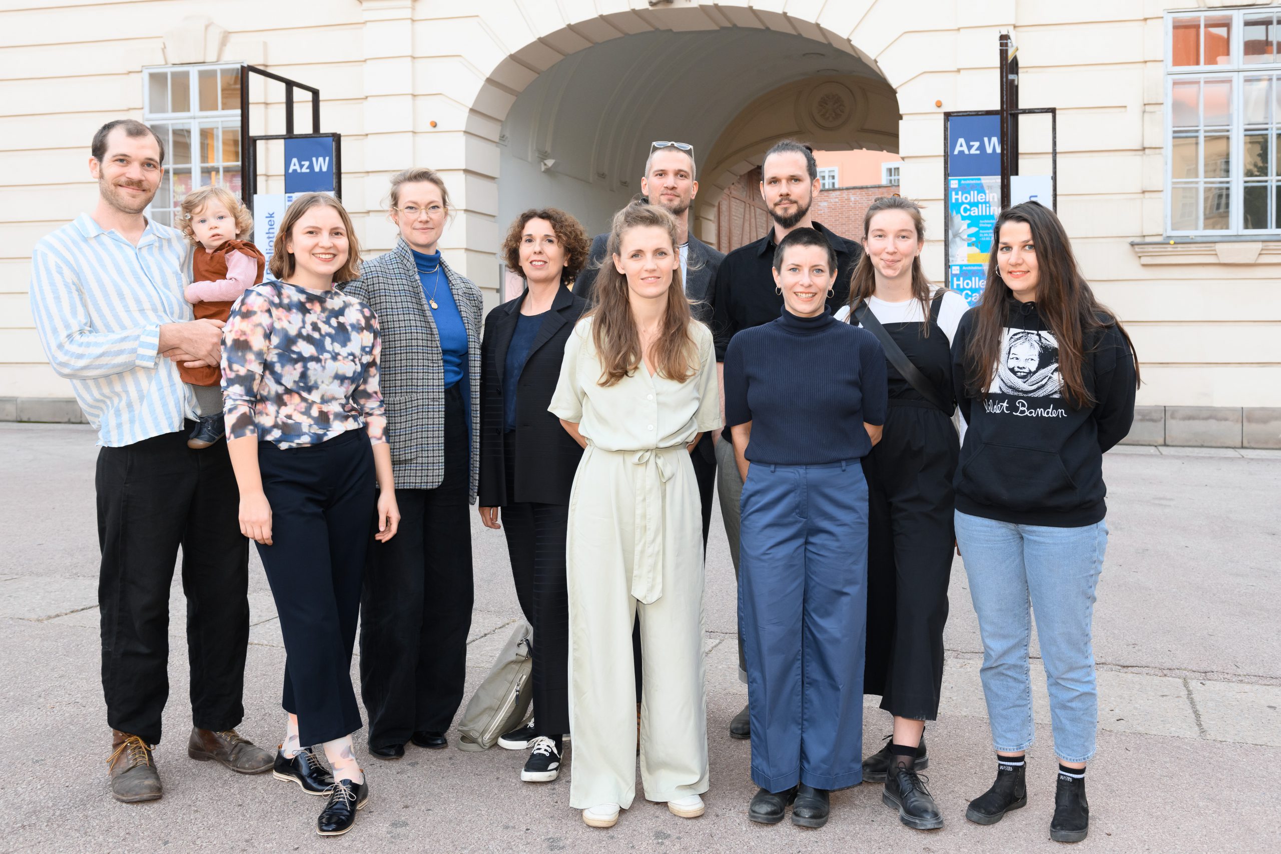 Several people standing in a courtyard, behind them posters and a blue square with Az W lettering