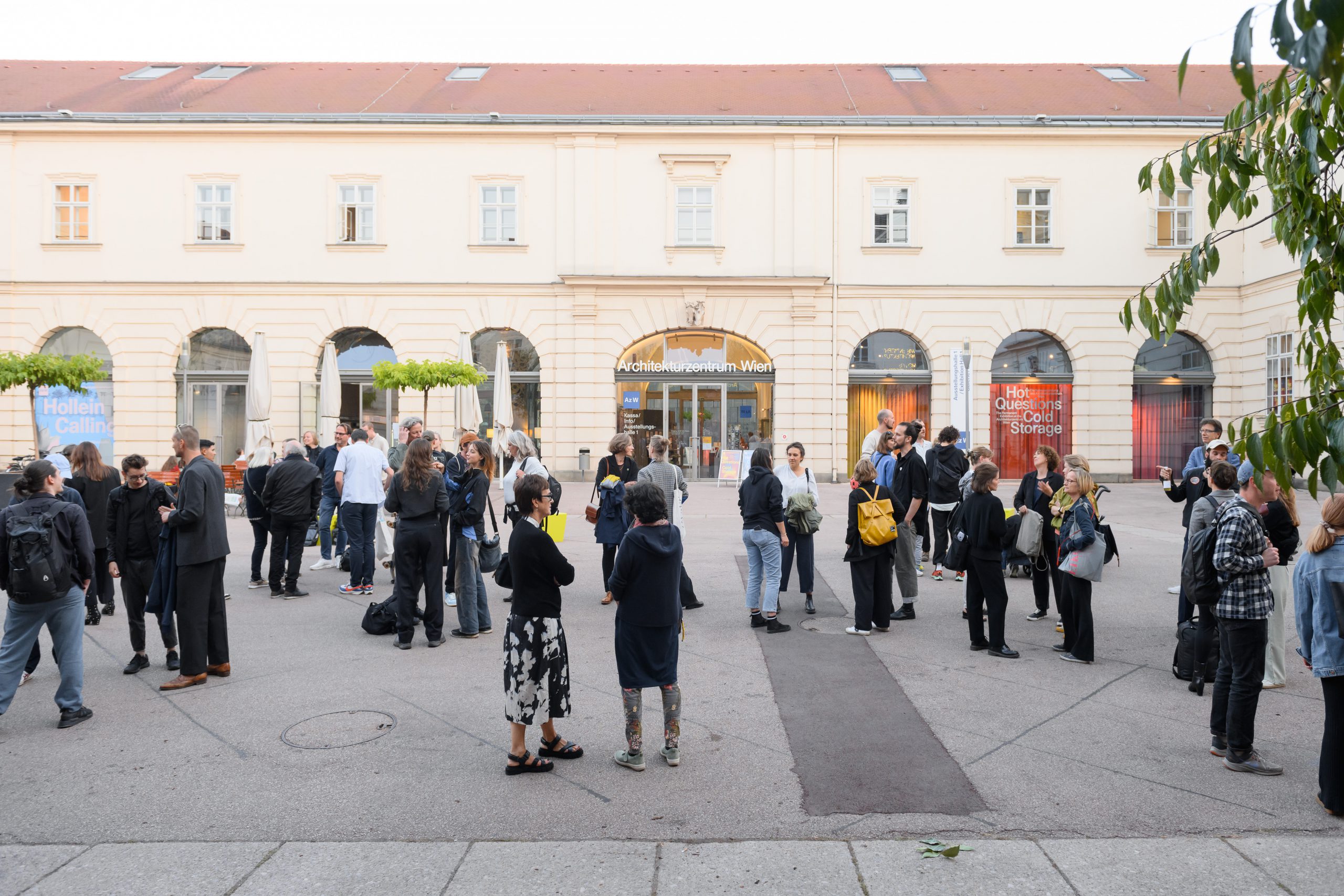 viele Menschen auf einem Platz mit ein paar Bäumen, dahinter eine große Tür darauf steht Architekturzentrum Wien