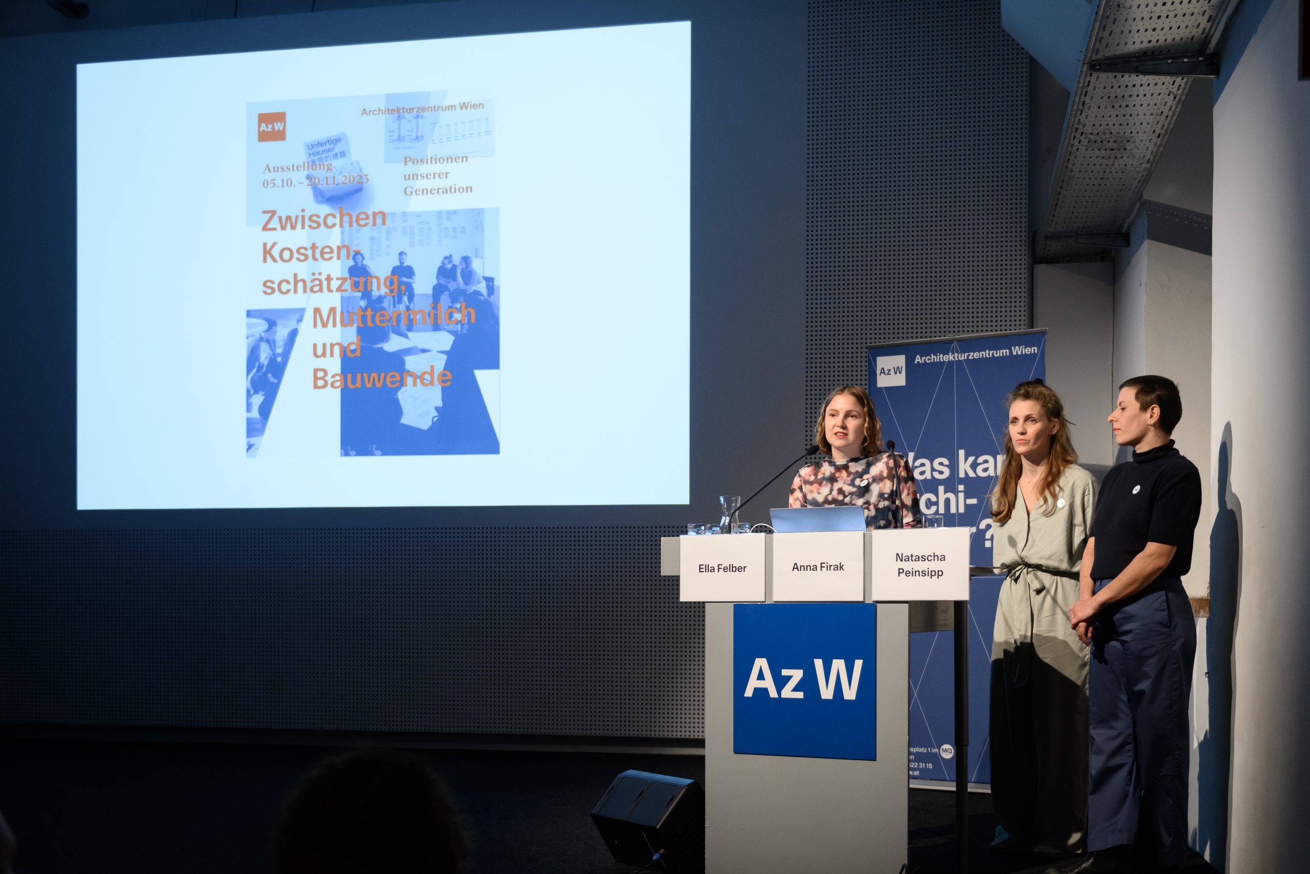 3 Frauen vor einem Rednerpult in einem abgedunkelten Raum, auf dem Rednerpult steht Az W auf blauem Quadrat