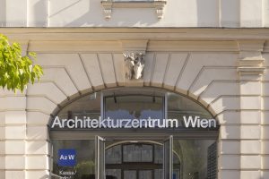 2 Frauem mit wallenden Röcken gehen vor dem Eingang mit Aufschrift Architekturzentrum Wien, aus dem Eingang kommt gerade ein Mann heraus der sich umdreht und nach hinten schaut