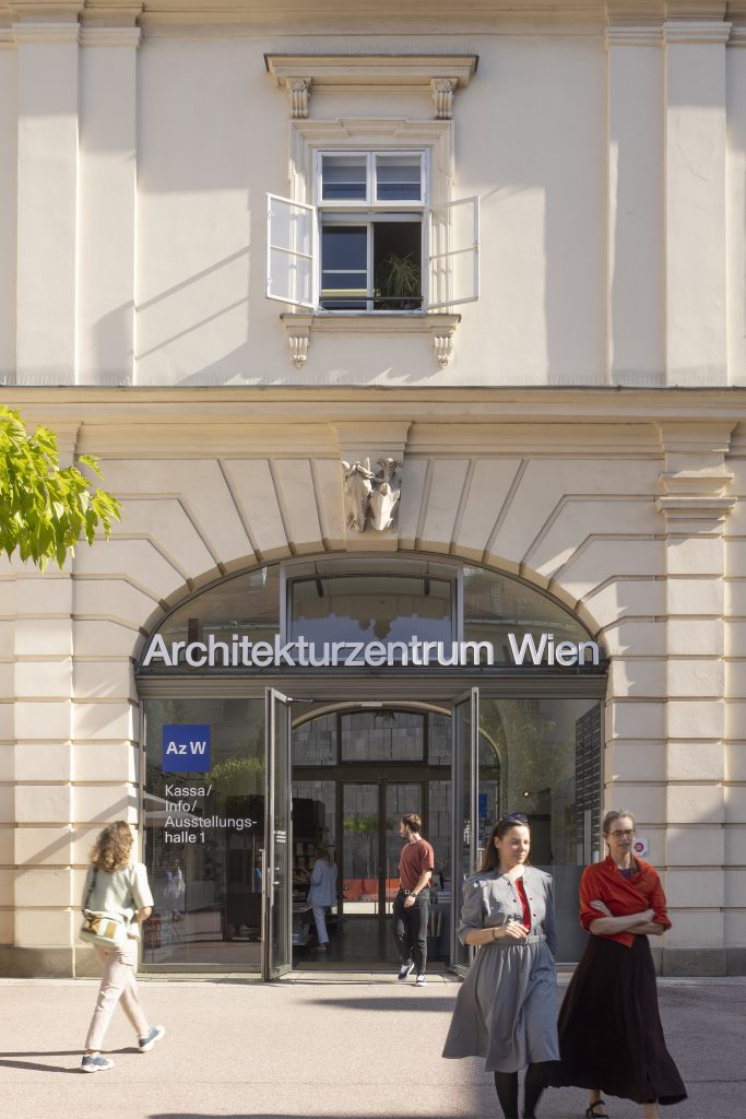 2 Frauem mit wallenden Röcken gehen vor dem Eingang mit Aufschrift Architekturzentrum Wien, aus dem Eingang kommt gerade ein Mann heraus der sich umdreht und nach hinten schaut