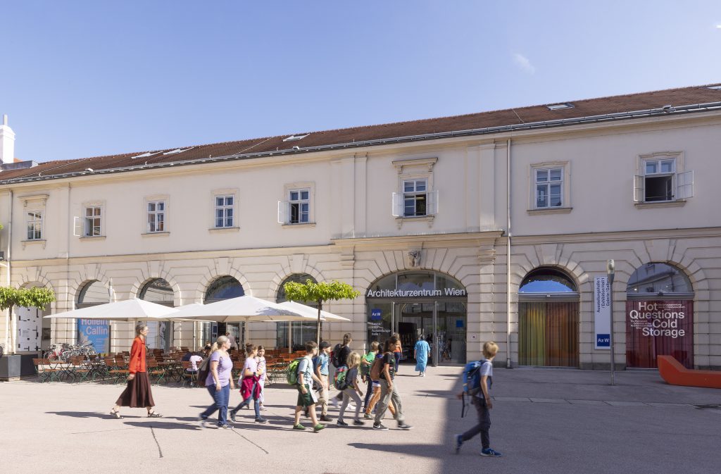 eine Gruppe von Menschen geht über einen Platz im Sonnenschein, im Hintergrund sieht man ein altes Gebäude mit der Aufschrift Architekturzentrum Wien und daneben Gasthaustische und -stühle mit Sonnenschirmen