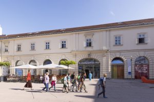 eine Gruppe von Menschen geht über einen Platz im Sonnenschein, im Hintergrund sieht man ein altes Gebäude mit der Aufschrift Architekturzentrum Wien und daneben Gasthaustische und -stühle mit Sonnenschirmen