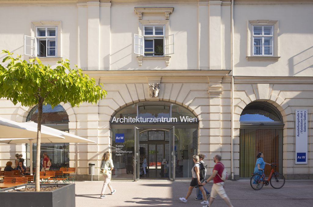 ein paar Menschen vor einem Eingang mit der Aufschrift Architekturzentrum Wien, daneben ein Baum in einem großen Topf und Sonnenschirme