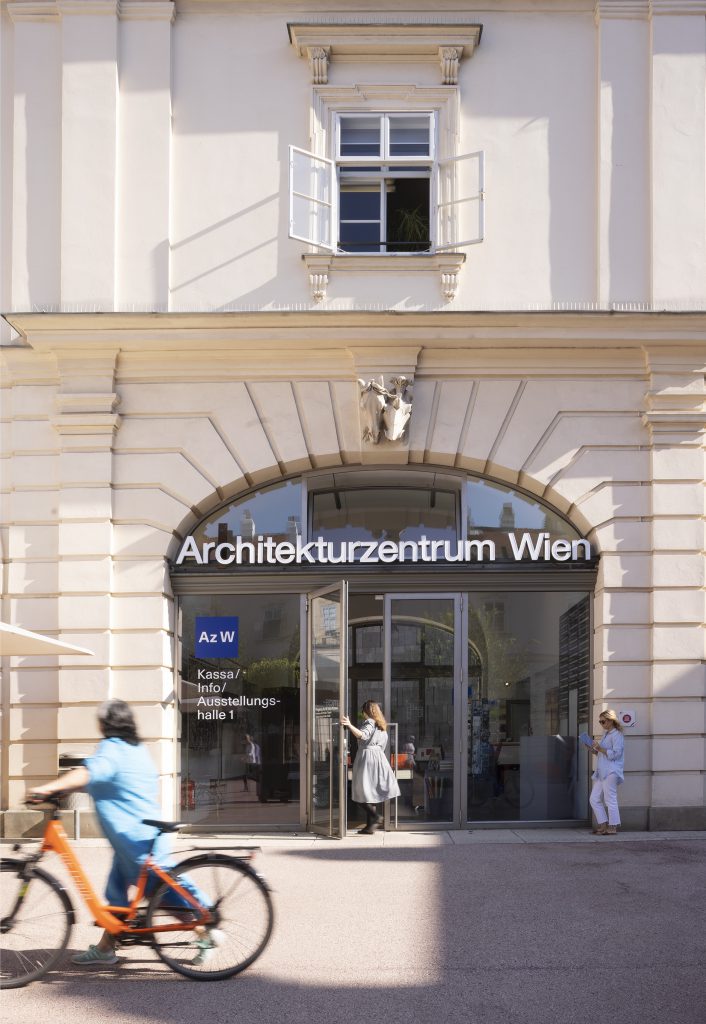 eine Frau mit hellblauem Kleid und orangefarbenen Fahrrad vor einem Eingang mit der Aufschrift Architekturzentrum Wien