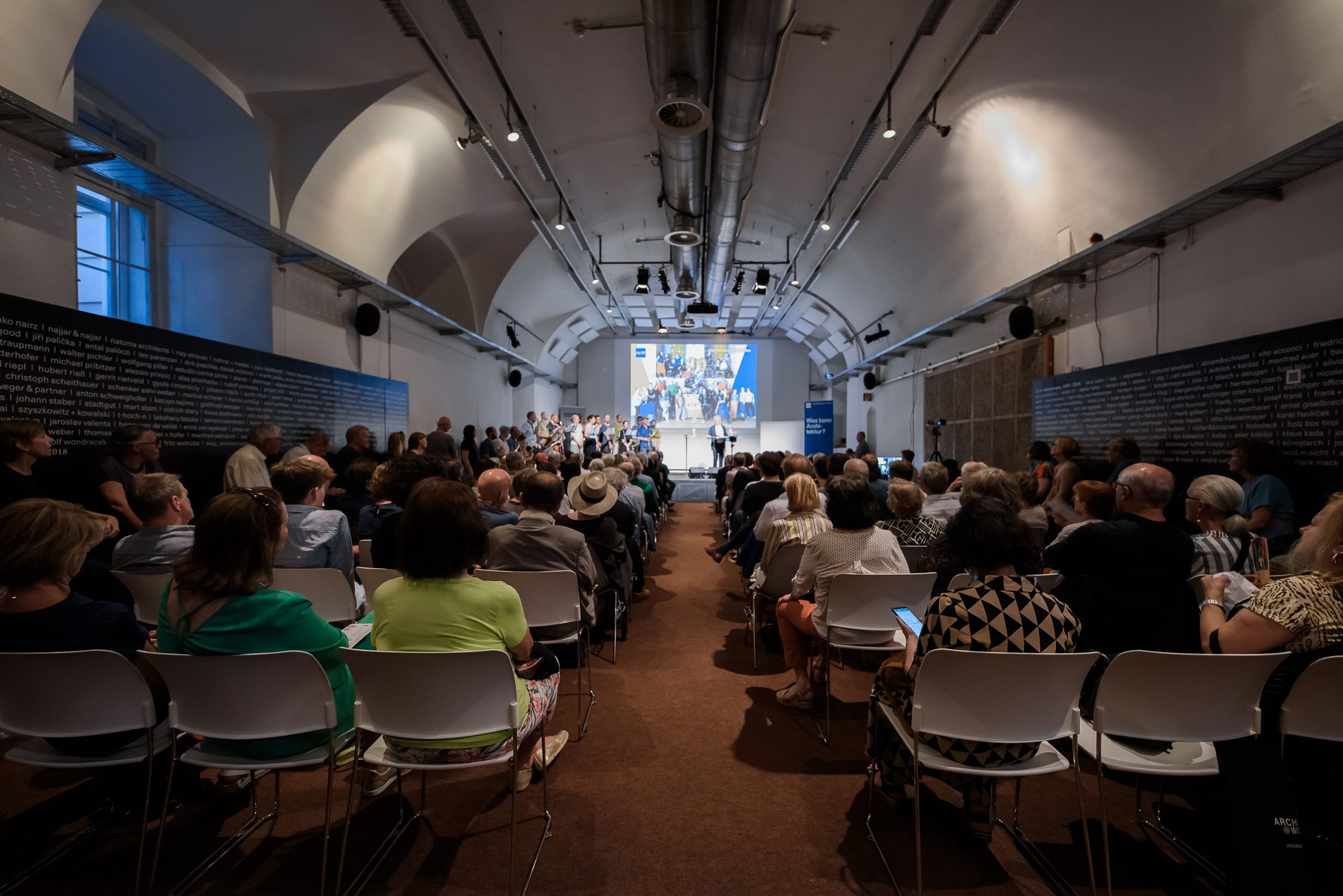 a room with many chairs in rows with people sitting on them, a screen with a projection and other people in the front