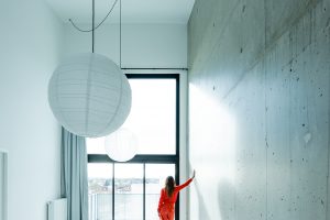 Woman with long hair and a red robe stands in front of a huge window and looks out