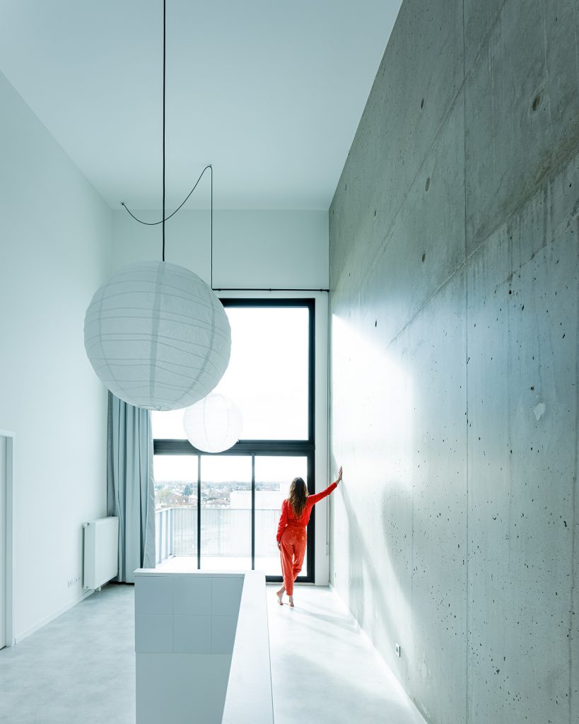 Woman with long hair and a red robe stands in front of a huge window and looks out