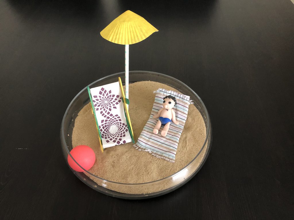 there is sand in a glass bowl and in it a small doll's deckchair and a small figure made of modelling clay lying on a tiny towel, next to it a small ball and a small parasol